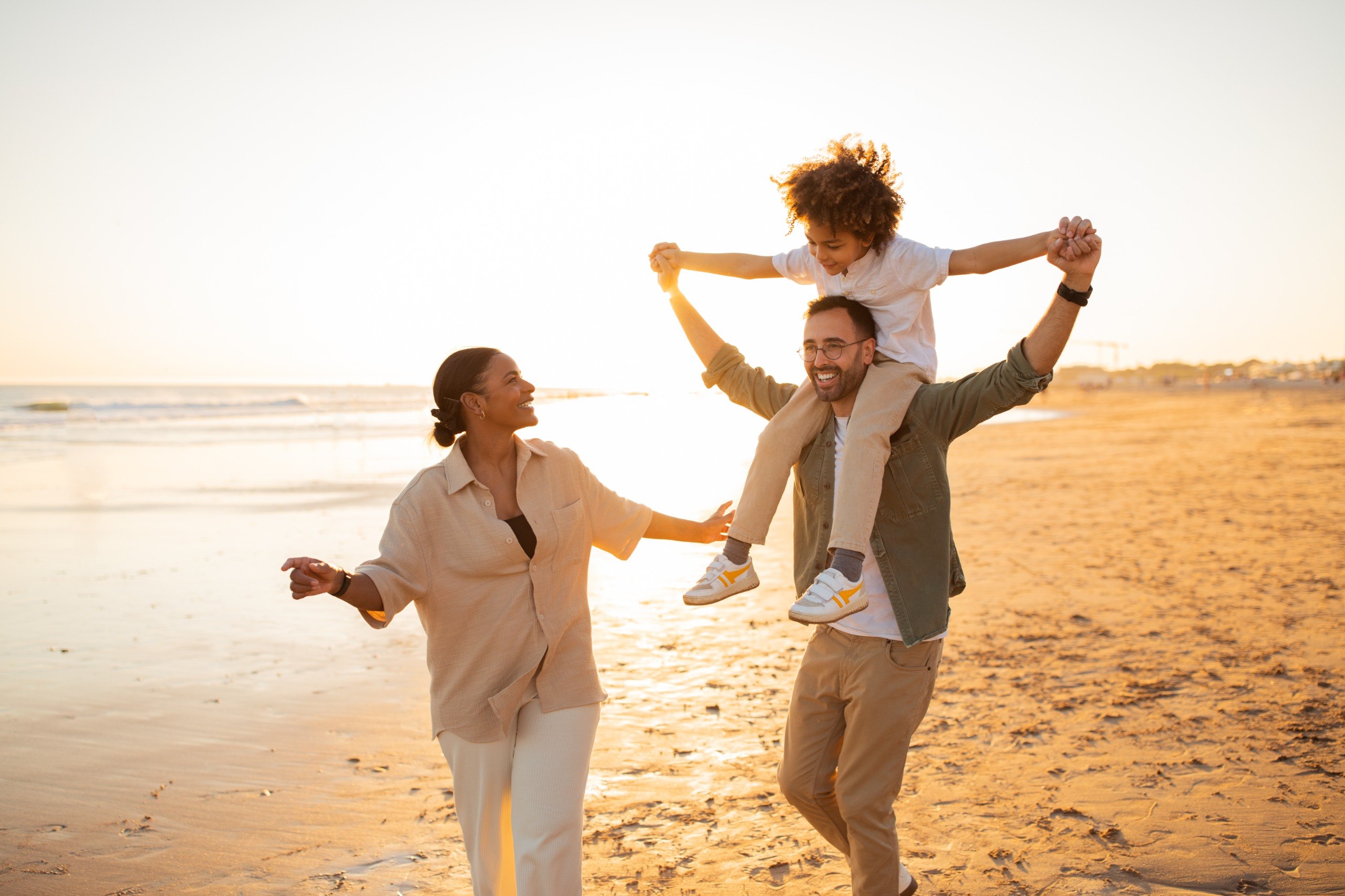 Familia feliz divirtiéndose durante las vacaciones, padre cargando a su hijo sobre los hombros y caminando juntos por la playa, disfrutando de un cálido día al aire libre